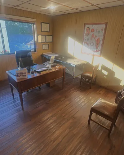 Consulta con paredes de madera: escritorio, camilla y lámina de la laringe, con sol entrando por la ventana.