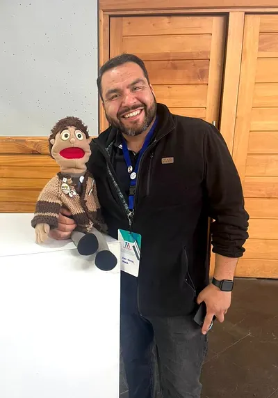 Juan Manuel Parada sonriendo junto a un títere de tela, con credencial colgada al cuello.